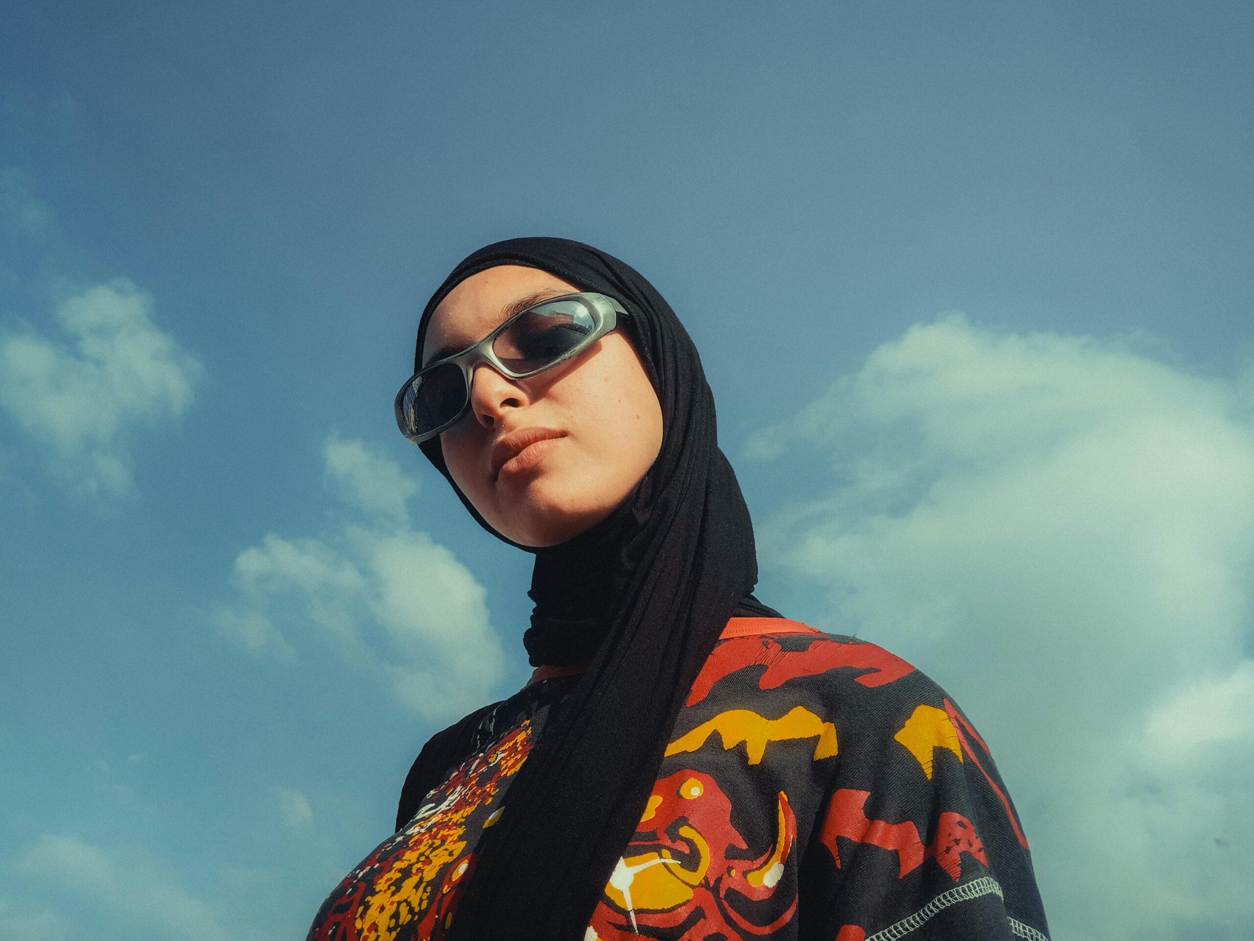 Stylish woman in sunglasses and hijab exuding confidence against a blue sky.