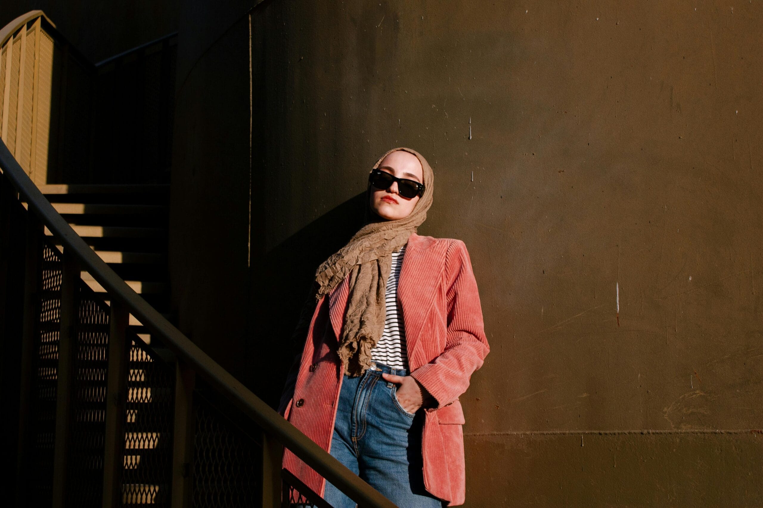 Fashionable woman wearing hijab and sunglasses posing by urban staircase, exuding confidence.